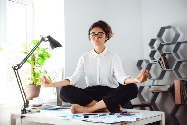 Young beautiful businesswoman in glasses meditating on table at workplace in office.